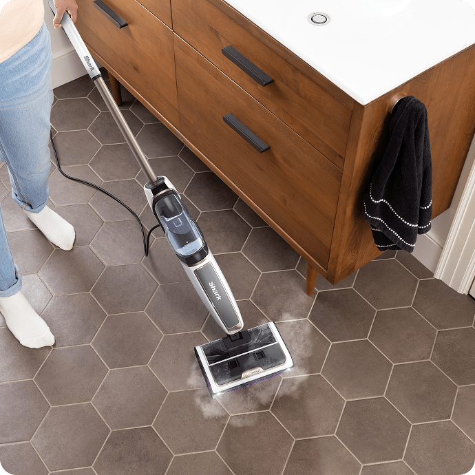 Person cleaning bathroom floor tiles with a Shark steam mop releasing steam